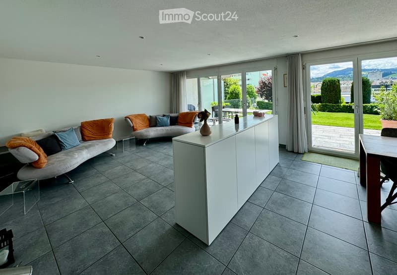 Modern living room with grey sofa, coffee table, and large windows leading to a patio. The space features a white kitchen island with a vase on top and a dining area with a table and chairs.