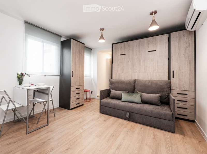 Modern, compact living space with a grey sofa, small dining table, folding chair, built-in cabinet, and decorative vase on the table.