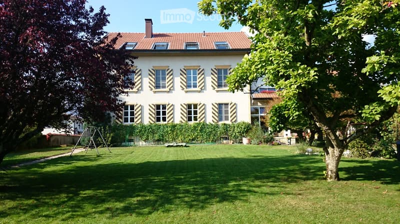 Two-story house with white walls, striped shutters, and a red-tiled roof. The house is surrounded by a well-maintained lawn, trees, and a small garden area with a swing set.