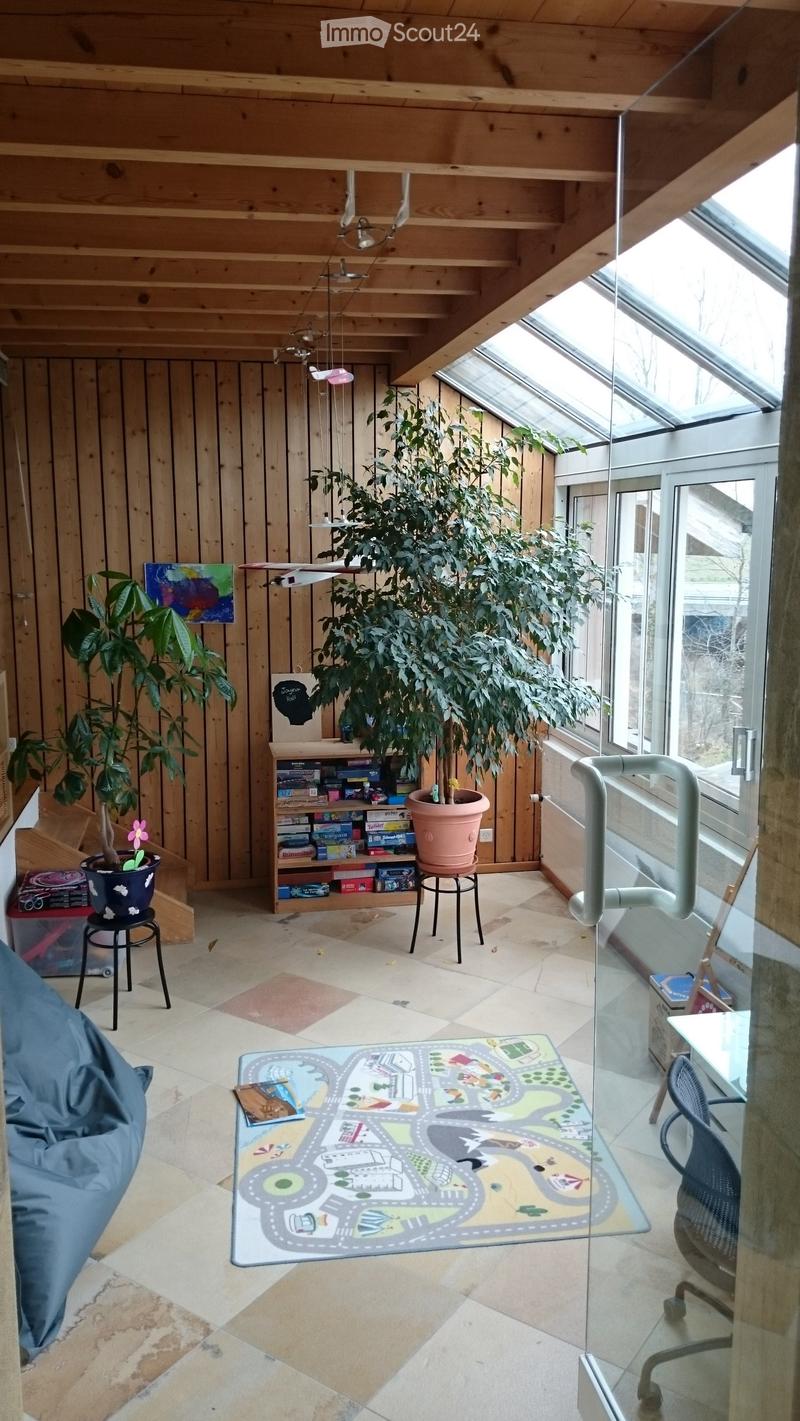 Bright, wooden-framed room with large windows and skylights, featuring a colorful play mat, a sofa, a shelf with toys and books, and several potted plants.