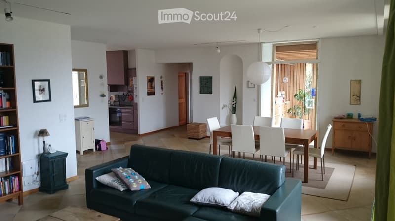Open-plan living and dining area with a green leather sofa, wooden dining table with white chairs, bookshelf, kitchen area with cabinets and oven, and decorative items including a vase and mirror.