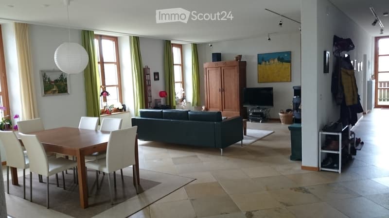 Bright living and dining area with a dark green sofa, wooden dining table with white chairs, large windows with green curtains, and a wooden cabinet.