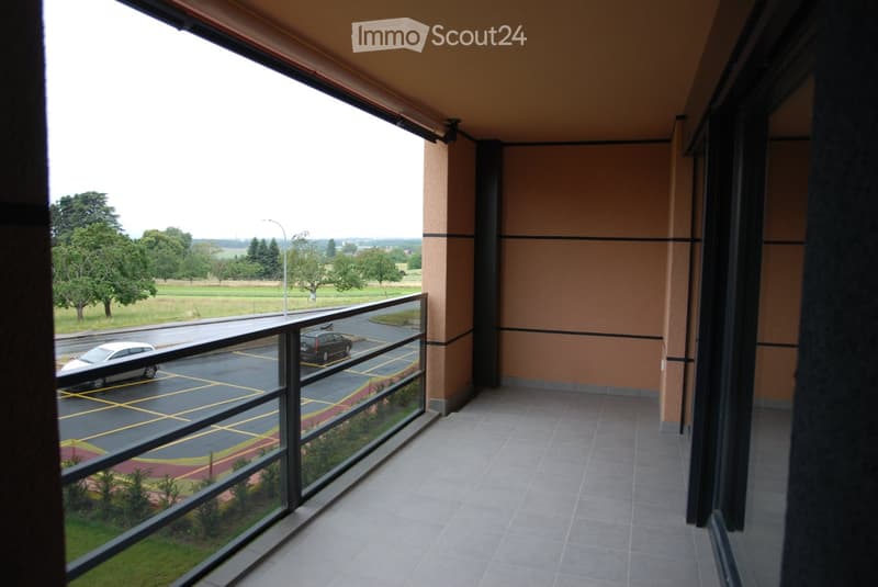 Balcony with large glass windows overlooking a parking lot and open field with trees.
