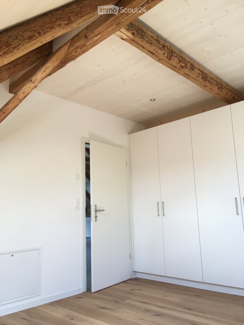 An unfurnished attic room with exposed wooden beams, white walls, and white built-in cabinets. The floor is made of light wooden planks.