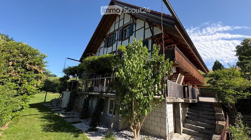 Traditional two-story house with wooden beams and white walls, featuring a balcony, staircase, and surrounding greenery.