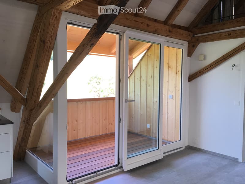 Open sliding glass doors lead to a wooden deck area with a wooden railing, exposed wooden beams, and a modern interior with white walls and grey flooring.