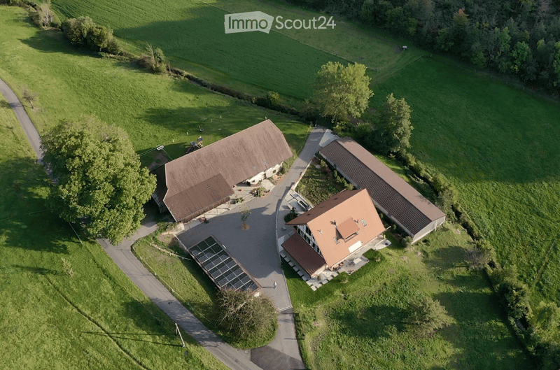 Aerial view of a two-story house with a solar panel installation on the roof, surrounded by green fields and trees.