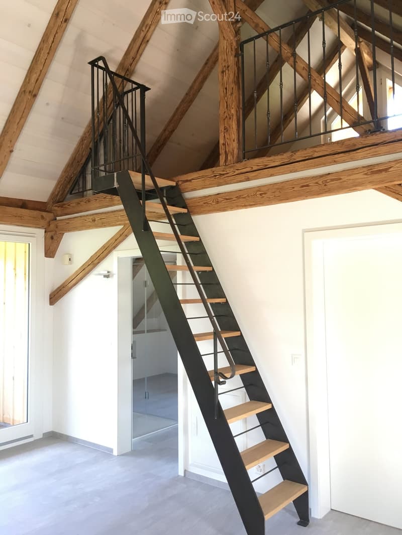 Modern hallway with exposed wooden beams and a sleek black metal staircase leading to an upper level. The staircase features wooden steps and a black railing, with a glass door visible in the background.