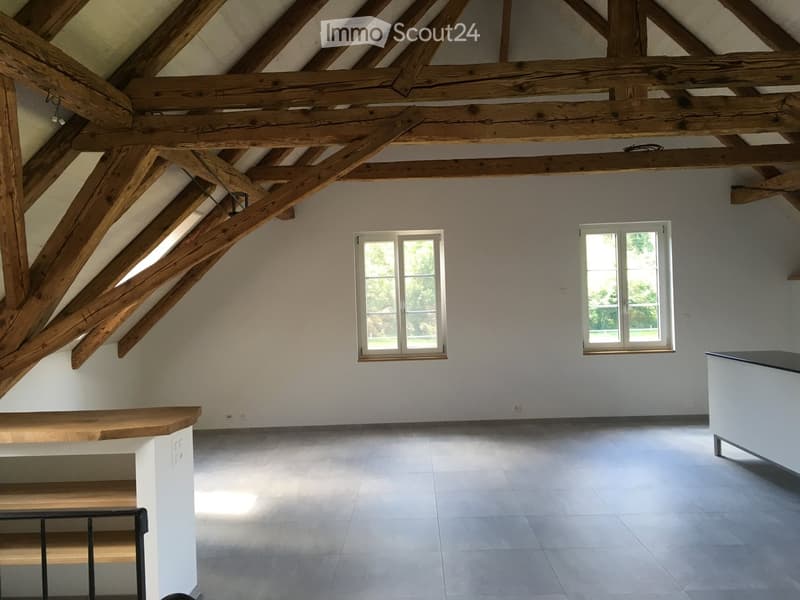 An empty attic space with exposed wooden beams, white walls, and large windows, featuring a wooden bench and a modern staircase railing.