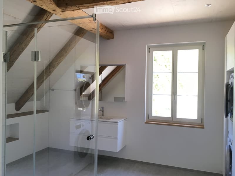Modern bathroom with a glass-enclosed shower, white sink, and large window letting in natural light. Exposed wooden beams on the ceiling add a rustic touch.