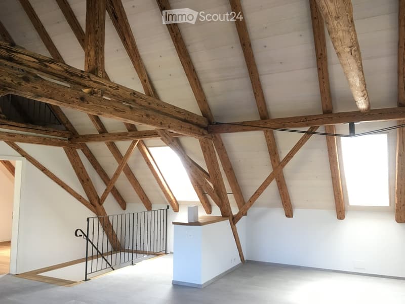 Open, high-ceilinged space with exposed wooden beams, white walls, and a small white counter. The room features a staircase with a black metal railing leading to an upper level.