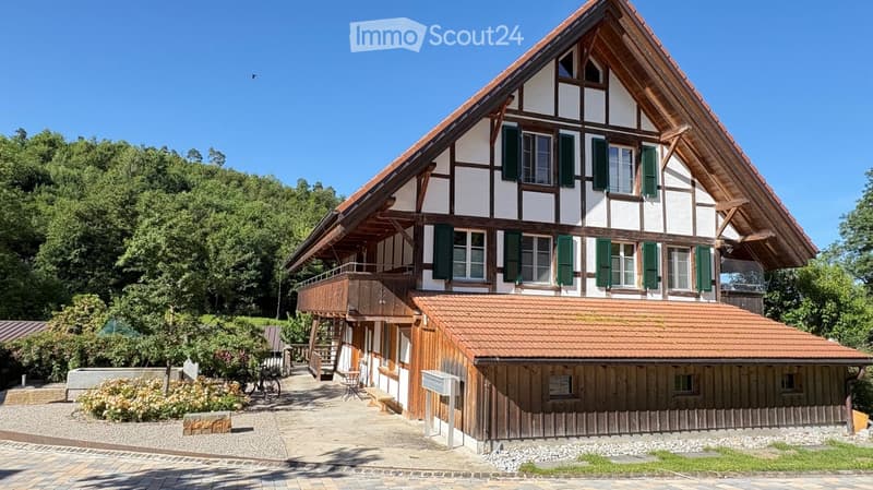 Traditional two-story house with white and brown timber framing, green shutters, and a wooden balcony. The house features a tiled roof and is surrounded by greenery and a paved pathway.