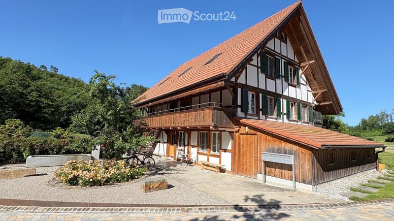 Traditional two-story house with a red-tiled roof, white and green shutters, wooden balcony, and attached wooden outbuilding.
