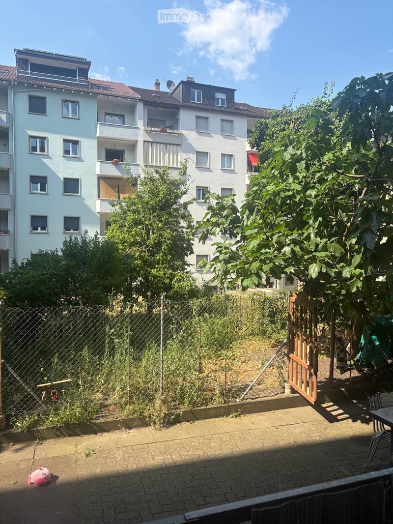 Garden area enclosed by a chain-link fence, with a wooden gate leading to a raised garden bed. The area includes a paved patio with a small table and chairs, a pink stuffed animal on the ground, and various plants and trees.