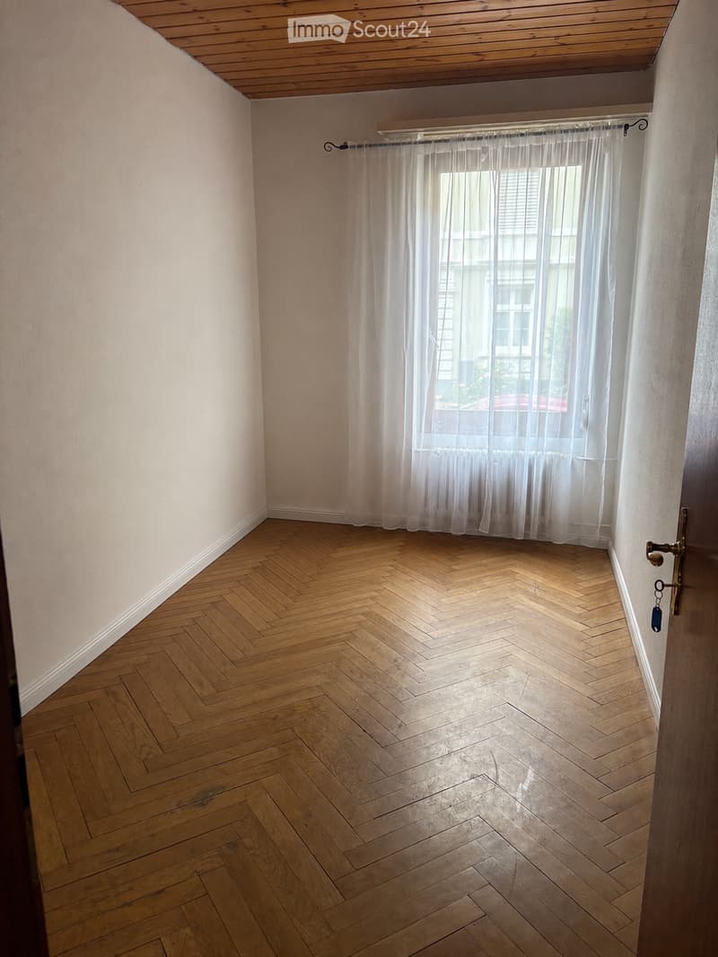 Empty room with wooden herringbone flooring, white walls, and a window with sheer curtains. The room has a simple, clean design with no furniture or decorations.