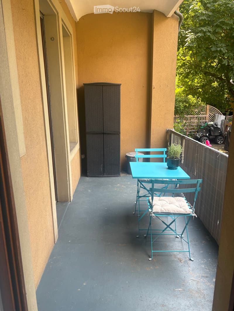 Balcony with a small turquoise table, two matching chairs, and a potted plant. The balcony has a dark grey floor and is enclosed by a wooden railing.