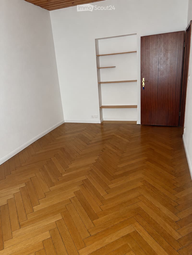 Empty room with wooden herringbone flooring, white walls, and a built-in wooden shelf unit. A closed wooden door is visible on the right side.