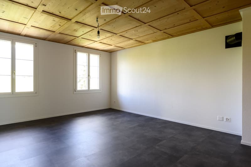 Empty room with white walls, dark grey tiled floor, and exposed wooden ceiling. Two windows with white frames provide natural light.