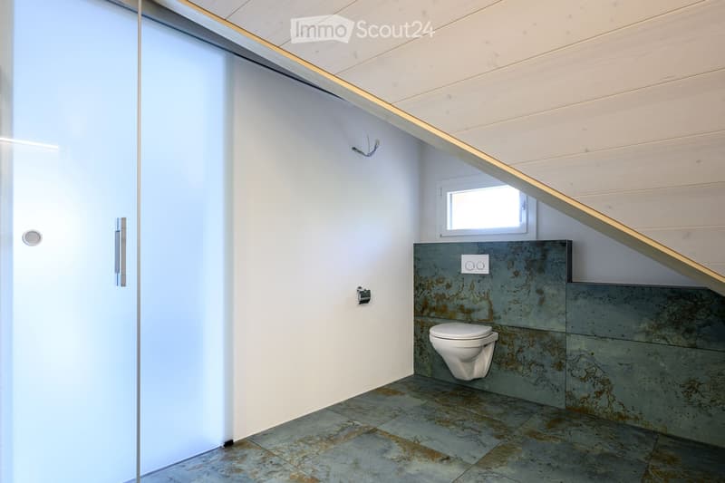 Modern bathroom with a white toilet, grey tiled walls, and a small window letting in natural light.