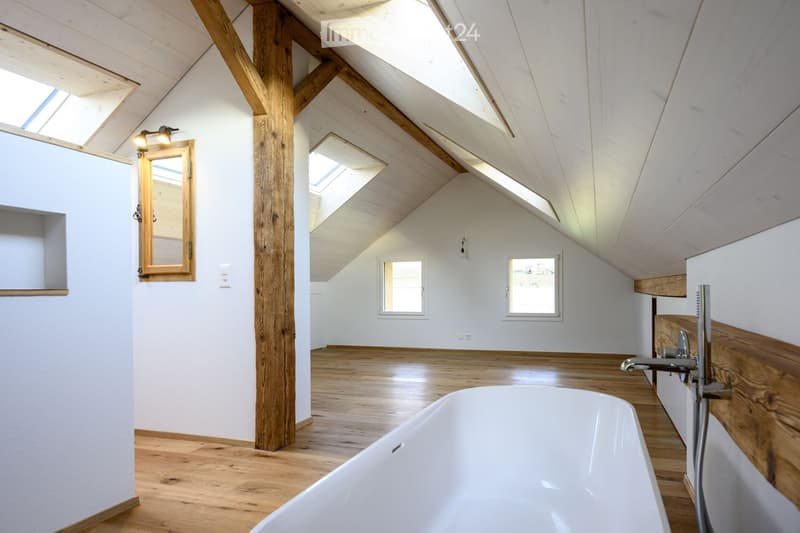 Bright, airy bathroom with wooden beams, skylights, and a freestanding bathtub. Features include a large mirror, wooden accents, and multiple windows allowing natural light.