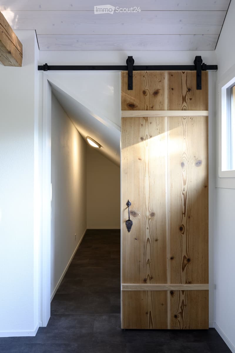 A modern hallway with a large wooden sliding door, white walls, and a dark floor. The door features a sleek black handle and a minimalist design.