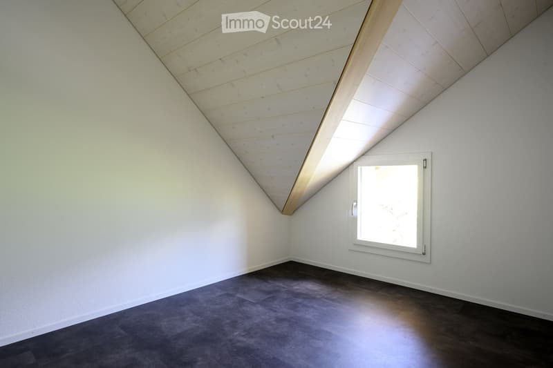 Empty room with wooden slatted ceiling, white walls, and a window letting in natural light. The floor is covered with a combination of dark wood and gray carpeting.