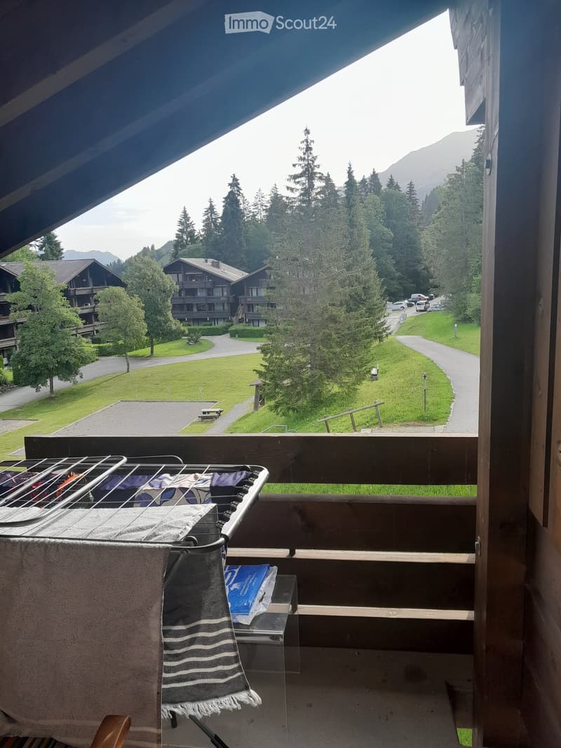 Balcony with a view of a green lawn, trees, and mountains. Includes a chair, table, and towel.