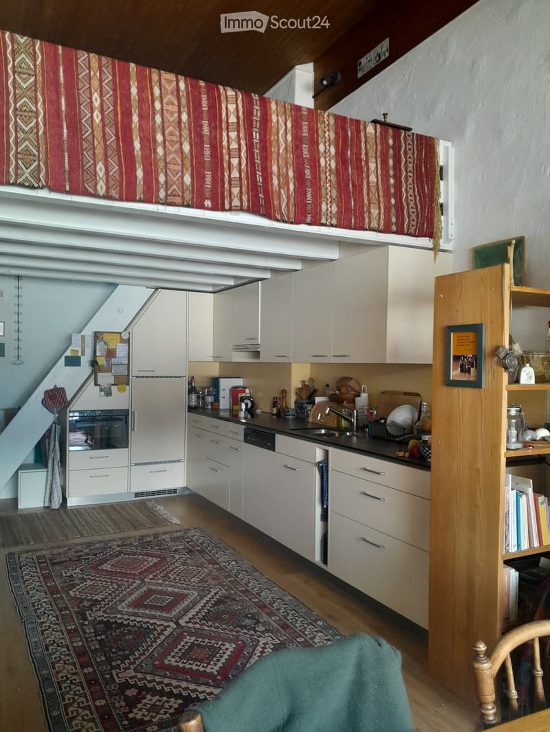 Modern kitchen with white cabinets, stainless steel appliances, wooden countertop, and a colorful rug on the floor. A small shelf with books and a framed picture is visible, along with a wooden chair and a blue cushion.