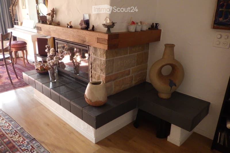 Living room with a stone fireplace, wooden mantel, decorative vases, a chair, and a small table with a mirror and picture frame on the windowsill.