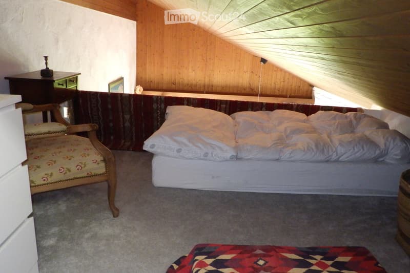 Cozy attic bedroom with wooden walls and slanted ceilings, featuring a double bed, a wooden armchair, a small table with a lamp, and a colorful rug on the floor.