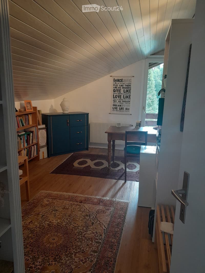 Cozy attic room with wooden floors, slanted ceilings, a desk, chair, bookshelf, cabinet, and decorative rugs.