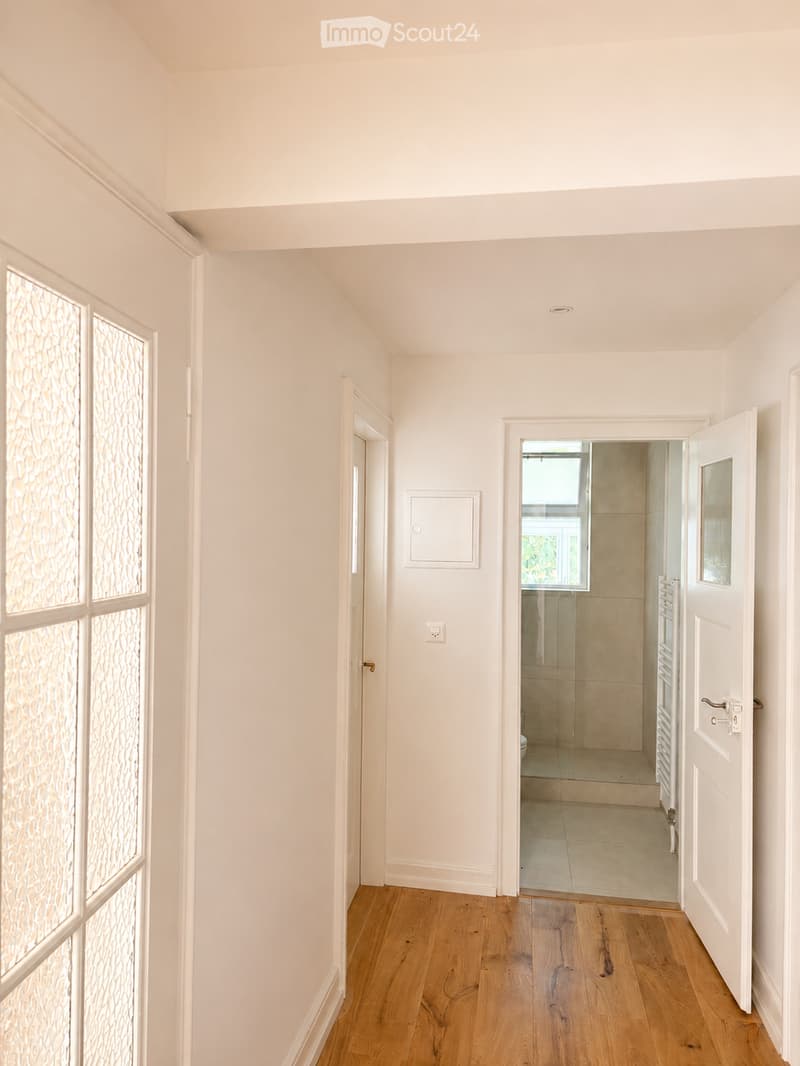Bright, minimalist hallway with light wooden flooring, white walls, and several doors leading to different rooms, including a bathroom with a glass shower enclosure visible through an open door.
