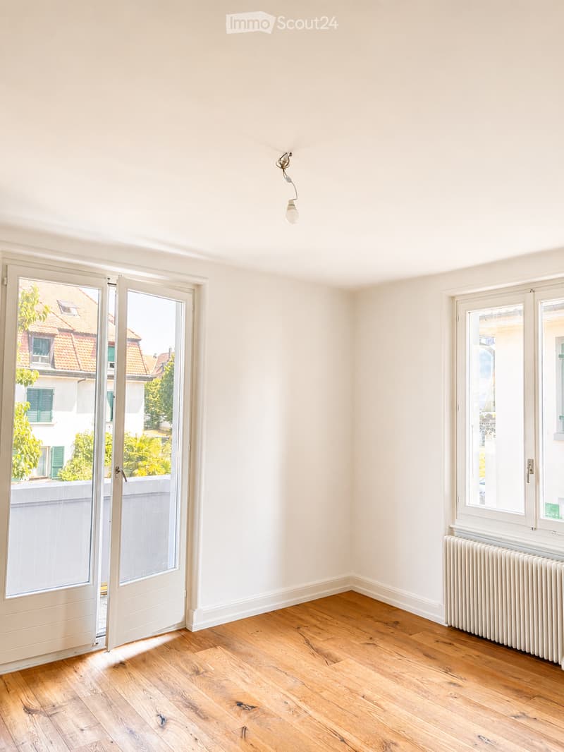 Empty room with wooden flooring, white walls, large windows, and a radiator. No furniture or equipment present.
