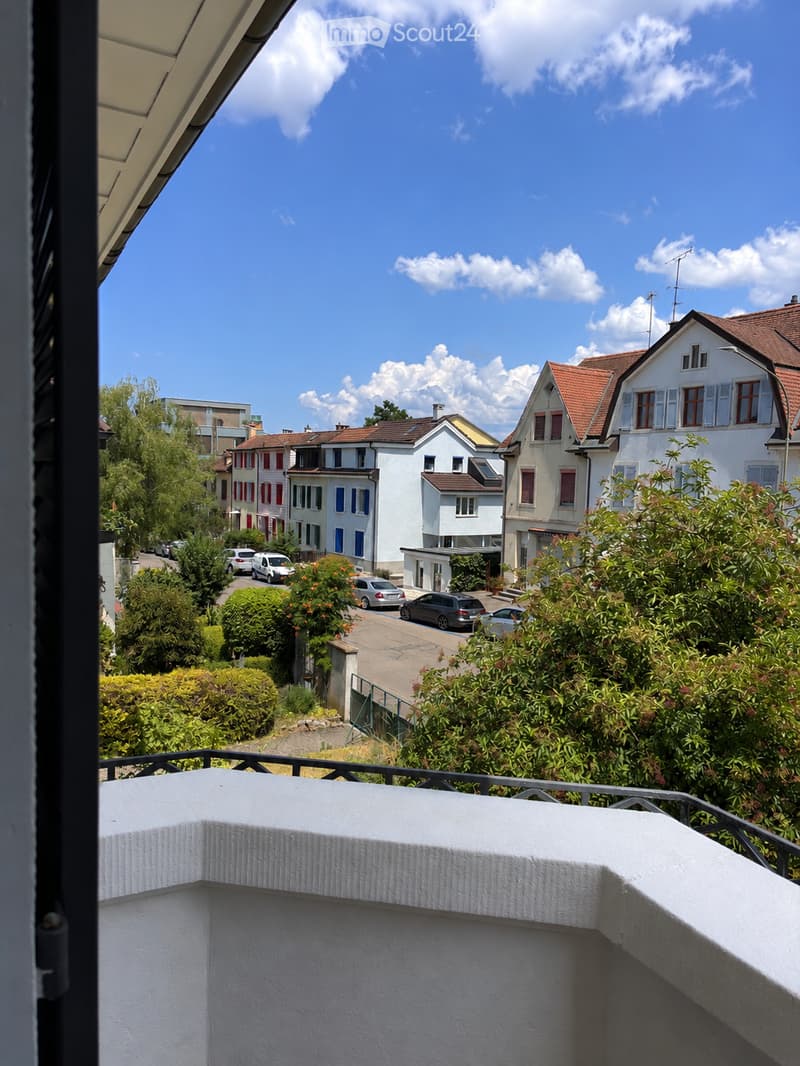 Balcony overlooking a residential street with houses, trees, and parked cars under a partly cloudy sky.