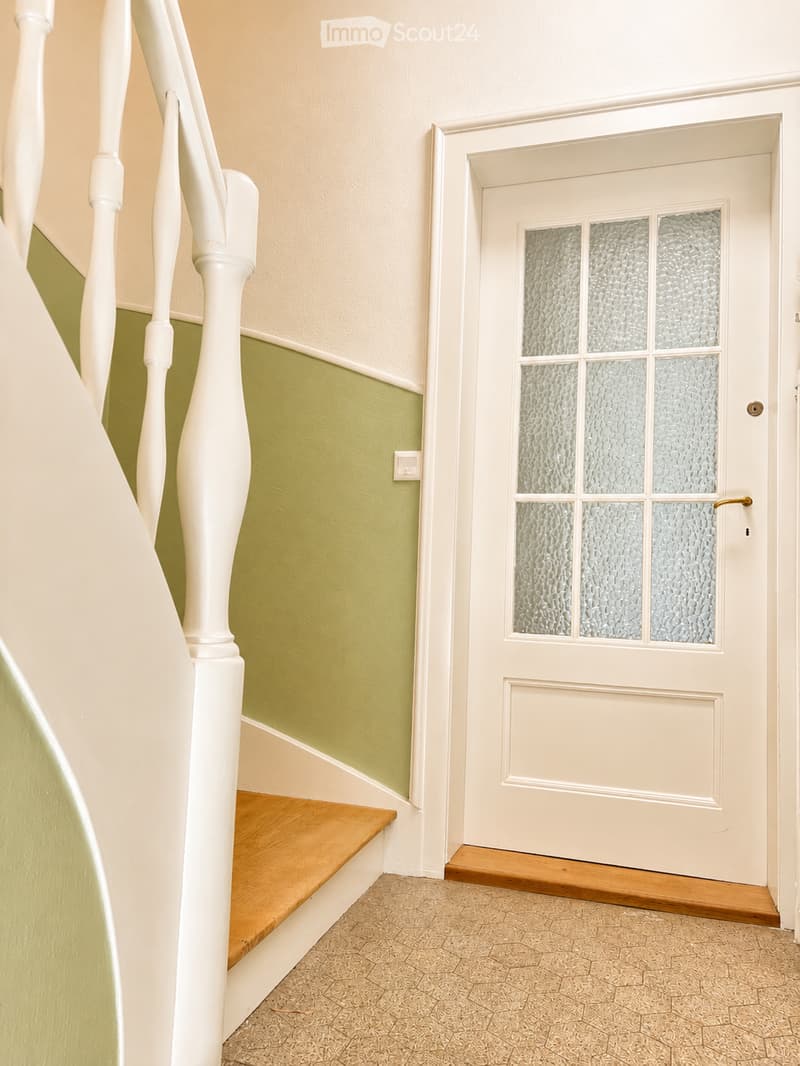 A hallway with a white door featuring frosted glass panels, a wooden floor, and a staircase with white balusters and a wooden handrail.