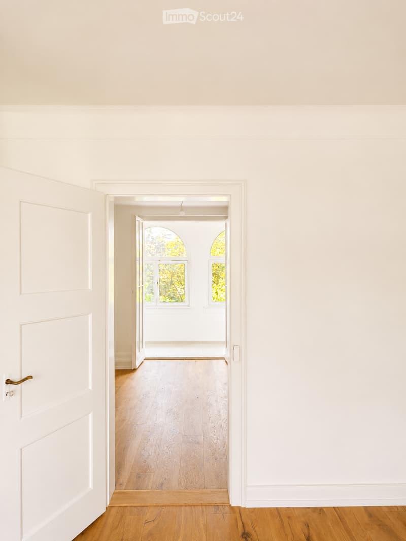 Empty hallway with light wooden flooring, white walls, and a door leading to another room with arched windows showing greenery outside.