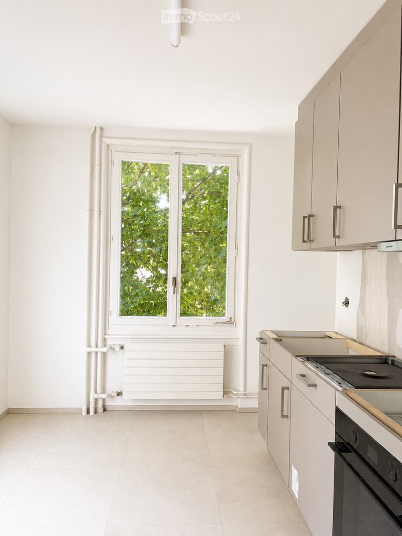 Modern, minimalist kitchen with white cabinets, a built-in oven, and a stove. Features a large window with a view of greenery, a radiator below the window, and a tiled floor.