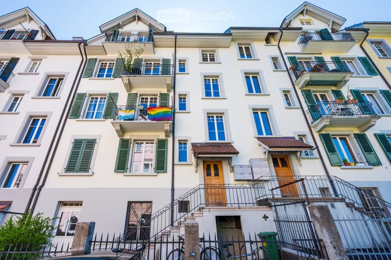 Multi-story residential building with white facade, green shutters, multiple balconies, and decorative flags on one balcony.