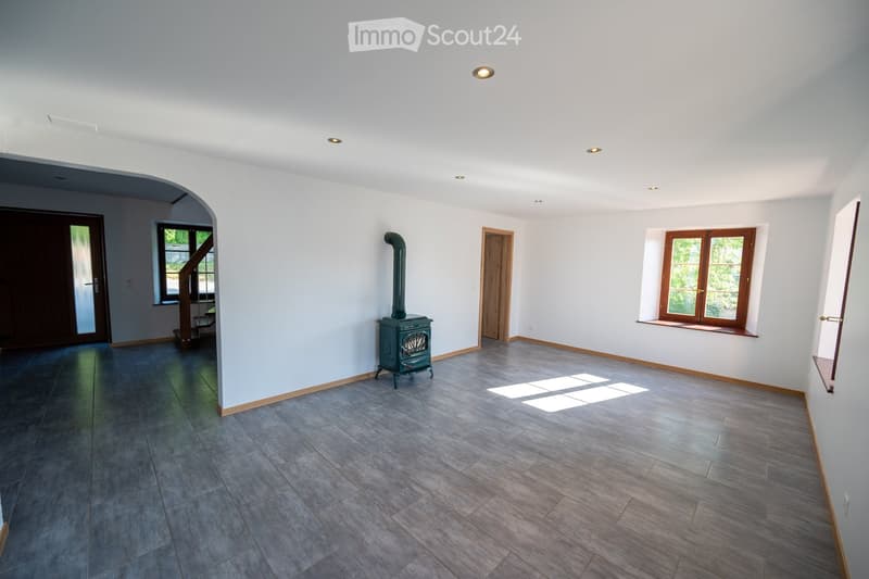 Empty room with light-colored walls, wooden flooring, and a fireplace in the center. The room has large windows and a door leading to a staircase.