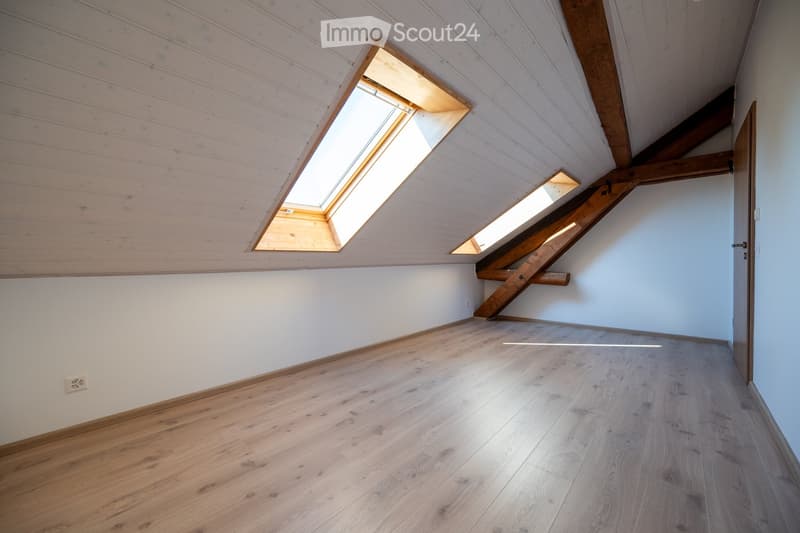 Empty attic room with wooden floors, white walls, exposed wooden beams, and skylight windows allowing natural light.