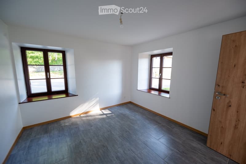 Empty room with grey tiled floor, white walls, and two windows with wooden frames and sills. Natural light streams in from the windows, and a wooden door is visible on the right side.