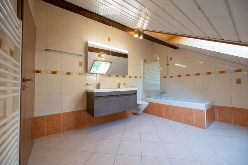 Modern bathroom with beige tiles, a large mirror, a sink with a modern faucet, a bathtub, and a toilet. The ceiling has exposed wooden beams and recessed lighting.