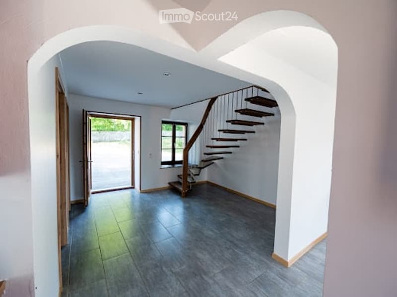 Open, modern hallway with a wooden staircase leading upwards, grey tiled floor, white walls, and large windows allowing natural light.