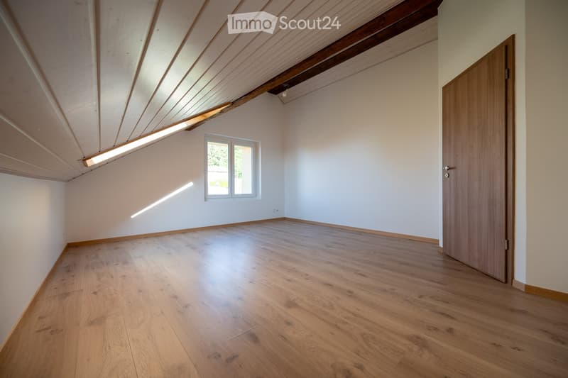 Empty room with wooden flooring, white walls, and a slanted wooden ceiling. Features a small window and a closed wooden door.