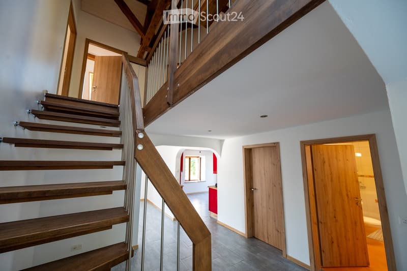 Spacious hallway with wooden staircase, metal railing, and multiple doors leading to different rooms.