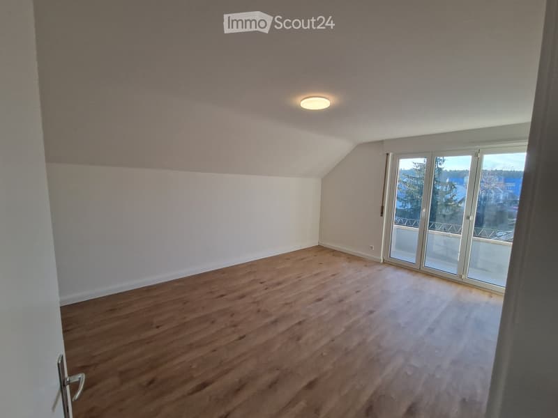 Empty room with wooden flooring, white walls, and large windows with a view of the outside. The room has a slanted ceiling and a single light fixture on the ceiling.