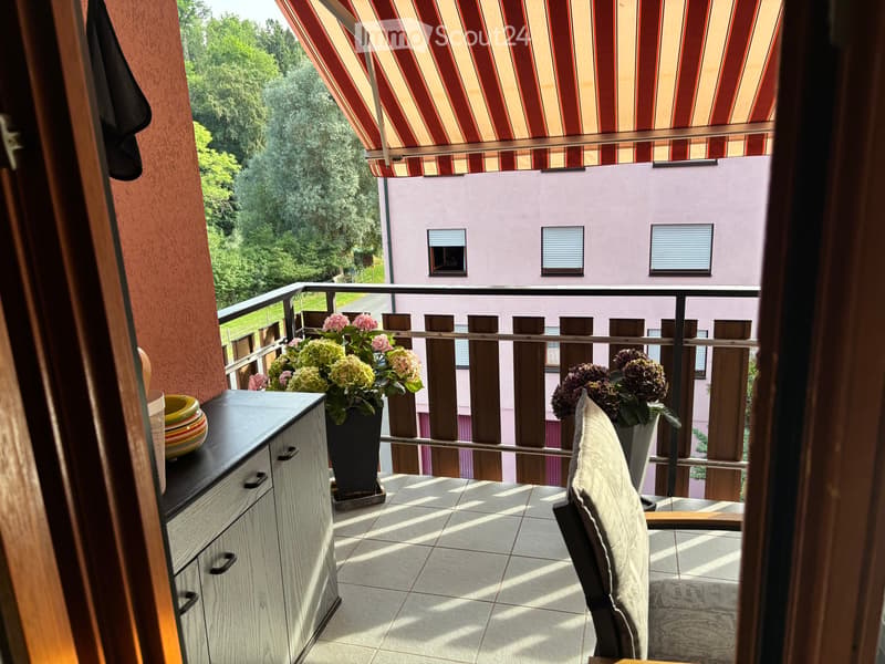 Balcony with a striped awning, tiled floor, wooden railing, and potted plants, featuring a small table and chair.