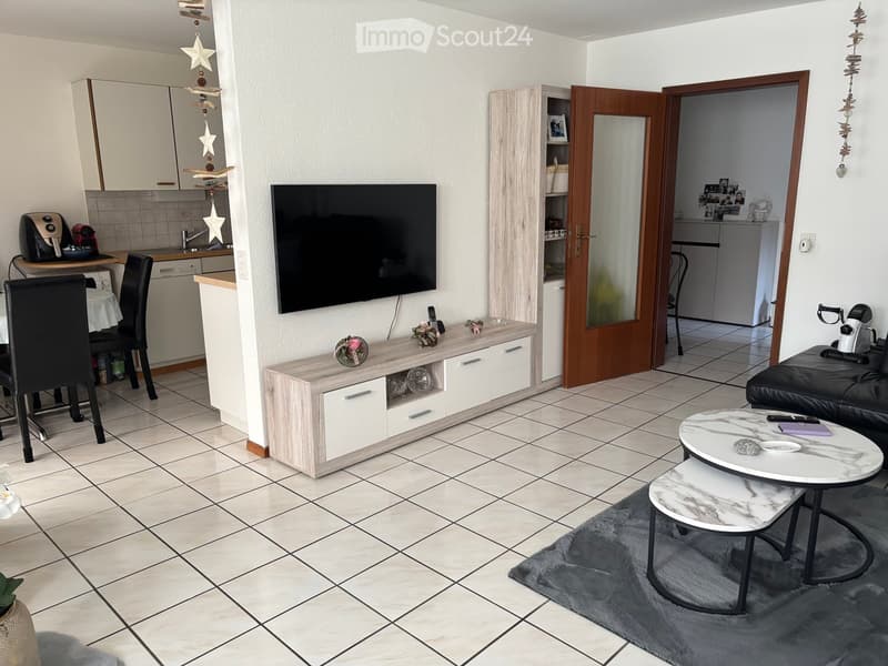 Modern living room with a large TV, a sofa, a coffee table, a dining area with a table and chairs, and an open kitchen area with cabinets, appliances, and a countertop.