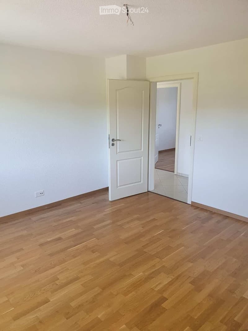 Empty room with light-colored wooden flooring and white walls. A white door is visible, leading to another room with a different flooring type.