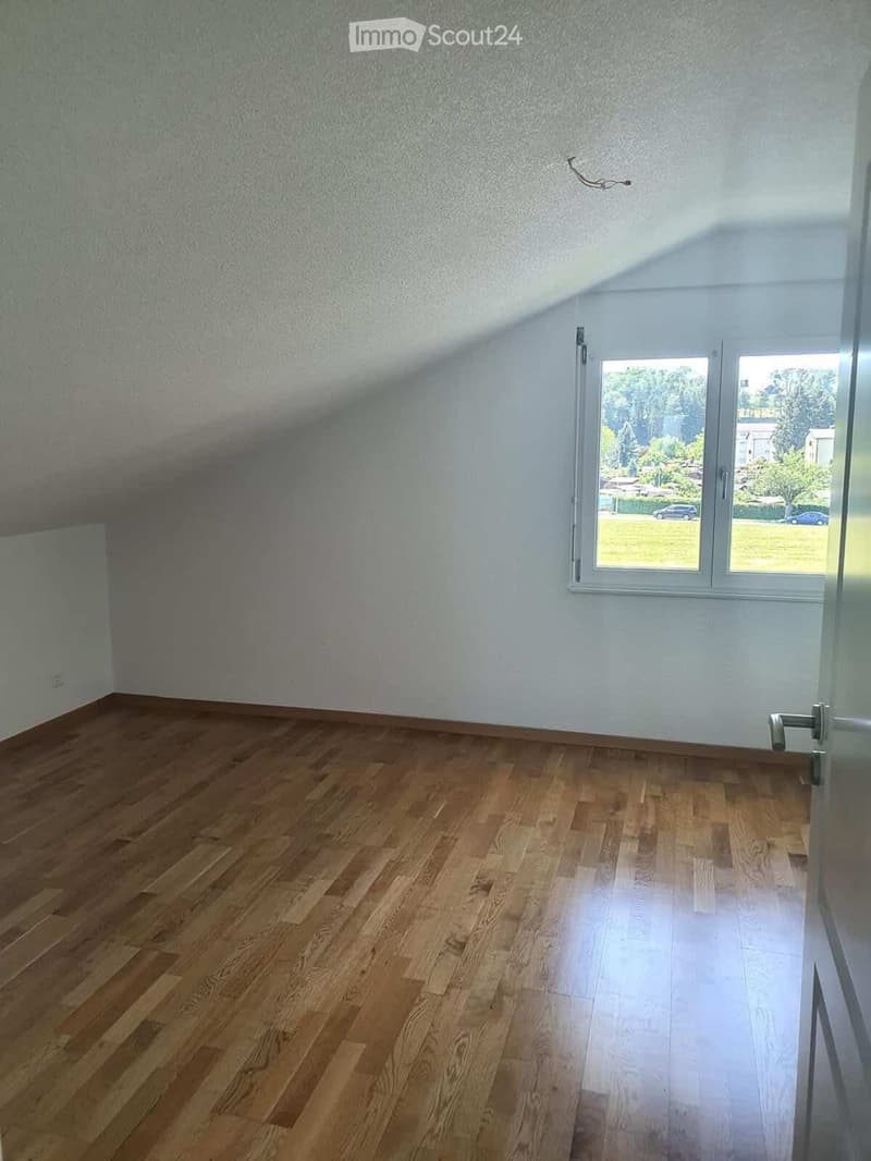 Empty room with wooden flooring and white walls, featuring a large window with a view of a green outdoor area.